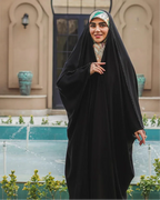 Smiling woman wearing black chador and patterned hijab standing near ornamental fountain in courtyard