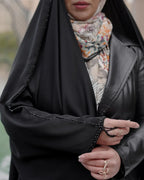 Woman wearing a black abaya with beaded trim and a colorful patterned scarf, showing gold rings on fingers